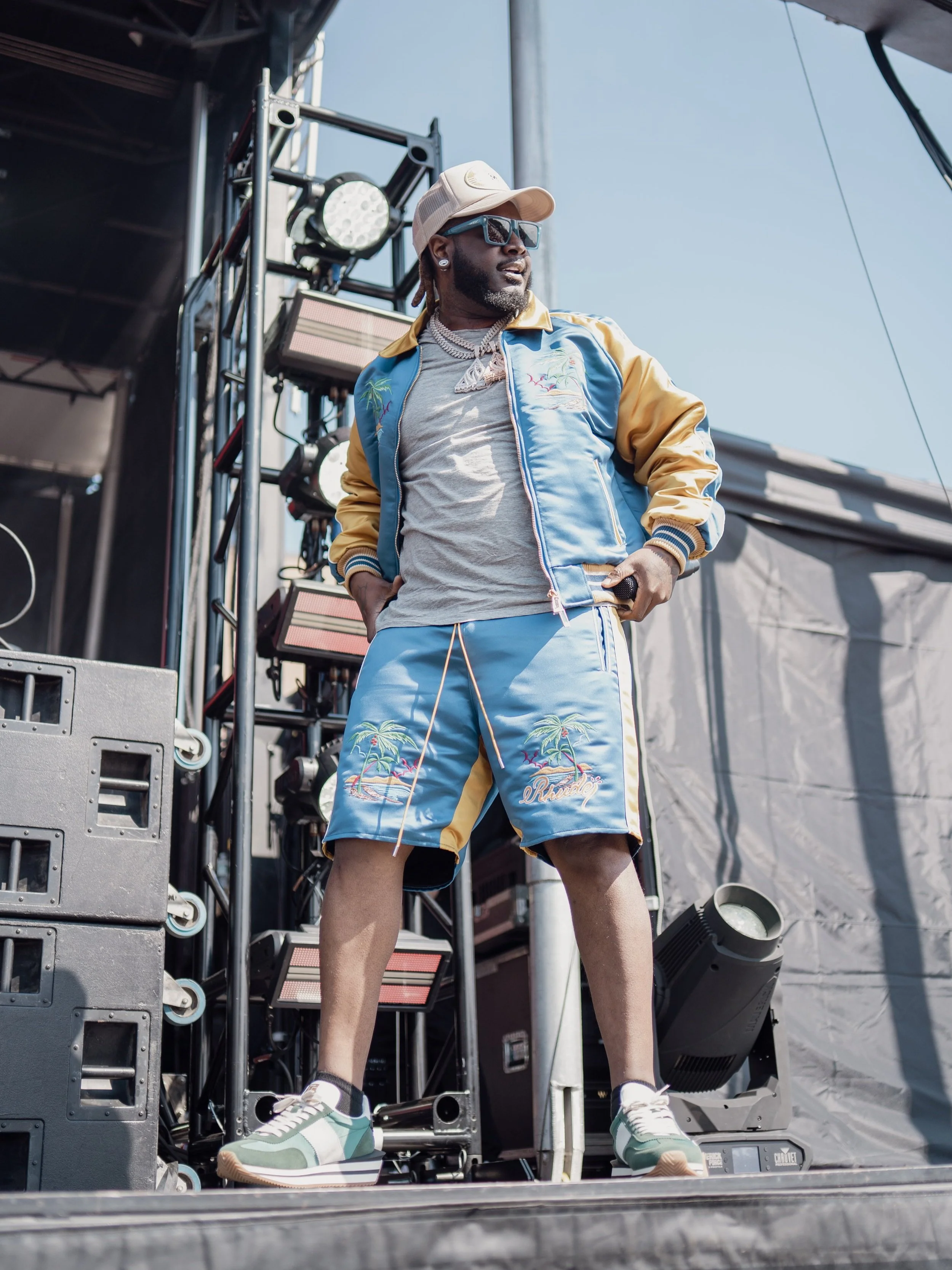 Man standing on stage during outdoor event, wearing sunglasses, baseball cap, colorful jacket and shorts, with music equipment in background.