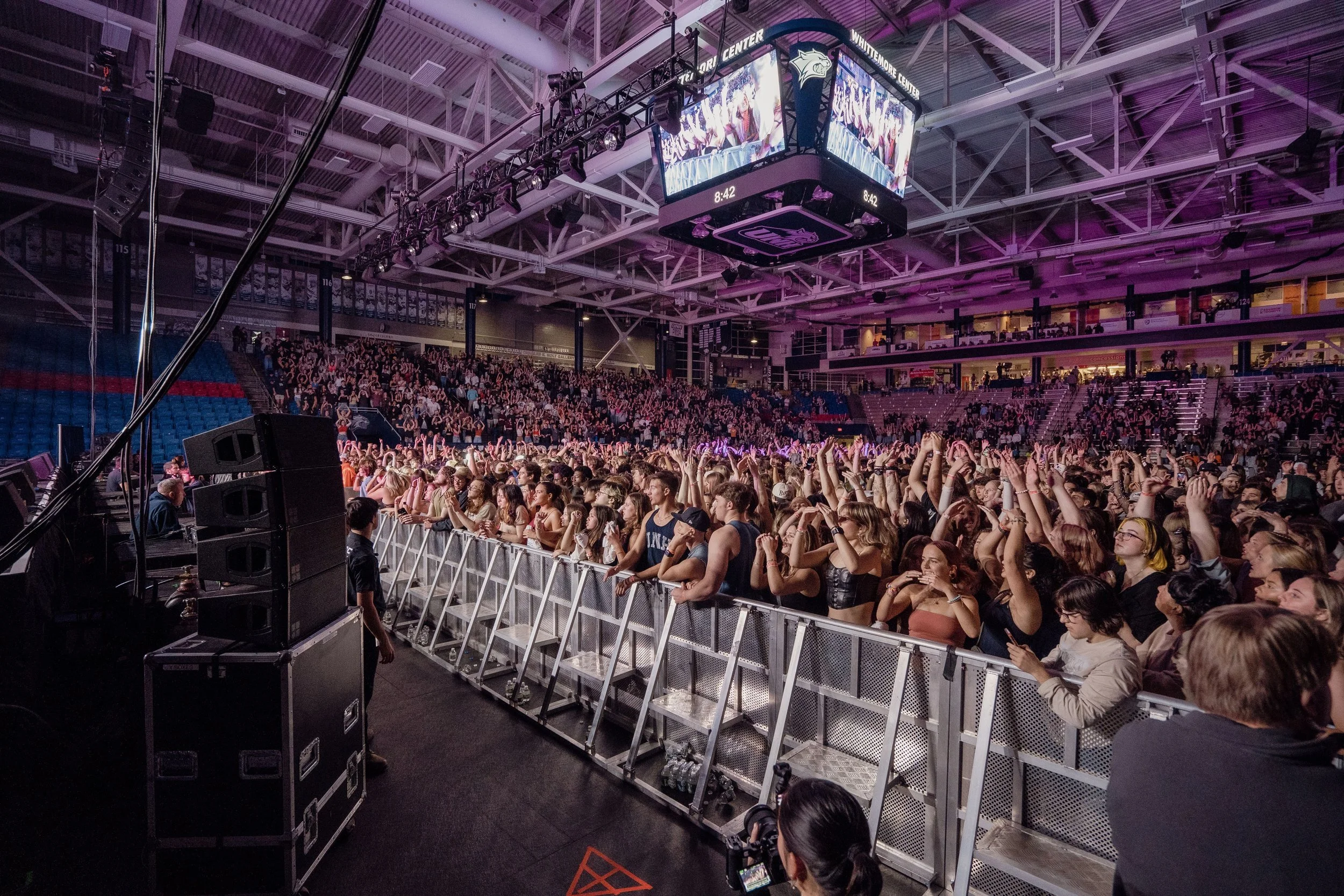 Crowd of concertgoers at the Whittemore Center arena at the University of New Hampshire, standing behind a metal barrier, with many raising their hands. The arena has a high ceiling with metal beams.