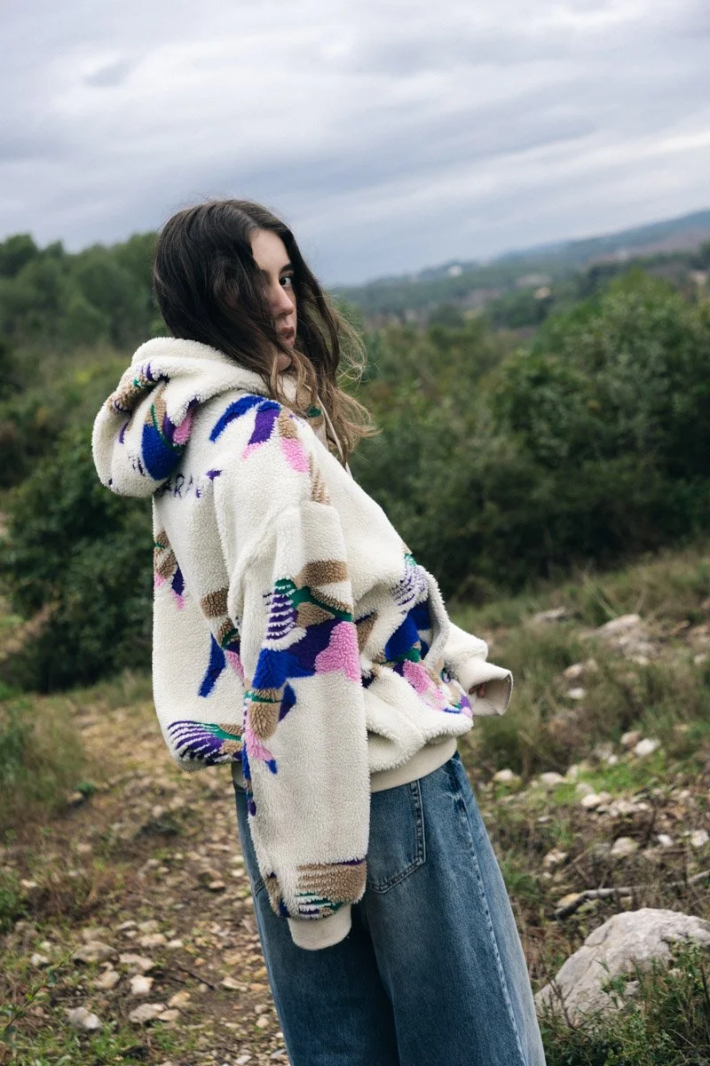 Young woman with long brown hair wearing a colorful fleece hoodie and blue jeans standing outdoors in a natural landscape with trees and cloudy sky in the background.