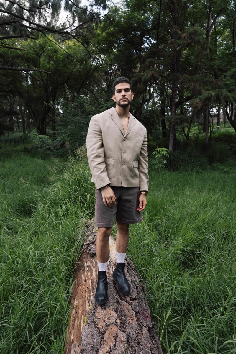 A young man standing barefoot on a fallen tree trunk in a grassy wooded area, wearing a beige blazer, black shorts, white socks, and black boots. Bernardo Flores photoshoot.