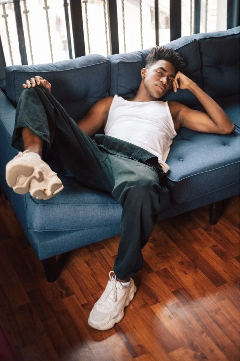Young man relaxing on a blue couch, lying with one leg bent and resting on the couch, wearing a white tank top, dark pants, and white sneakers.