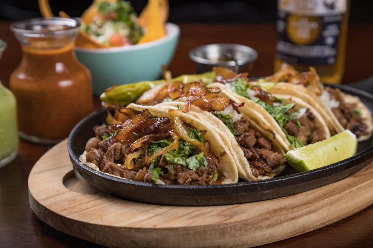 Three beef tacos with lettuce, onions, cilantro, and sauce, garnished with lime wedges, served on a sizzling platter with sides of salsa and beer in the background. Food photography in Mexico City