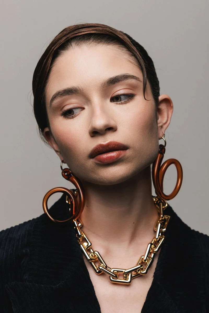 Close-up portrait of a woman with short dark hair, wearing large bronze hoop earrings and a chunky gold chain necklace, against a neutral gray background in a fashion photoshoot. By Sergio Gutiérrez sergiogutierrezphoto.
