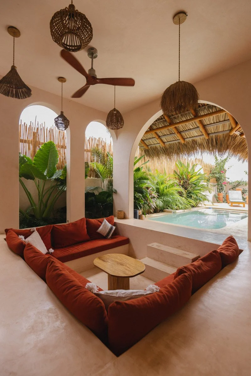 Indoor living area with a U-shaped orange sofa, small wooden table, ceiling fan, hanging wicker lamps, arched windows, lush tropical plants, and a view of a pool outside. Hotel Photography in Puerto Escondido Oaxaca Mexico. By sergiogutierrezphoto