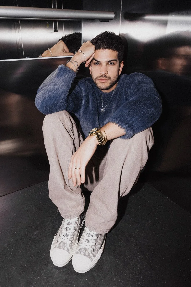 A young man with dark curly hair and a beard sitting on the floor in an elevator corner. He is wearing a dark blue fuzzy sweater, beige pants, and leopard-print sneakers. He has multiple jewelry pieces including a watch, bracelets, a ring, and a neck