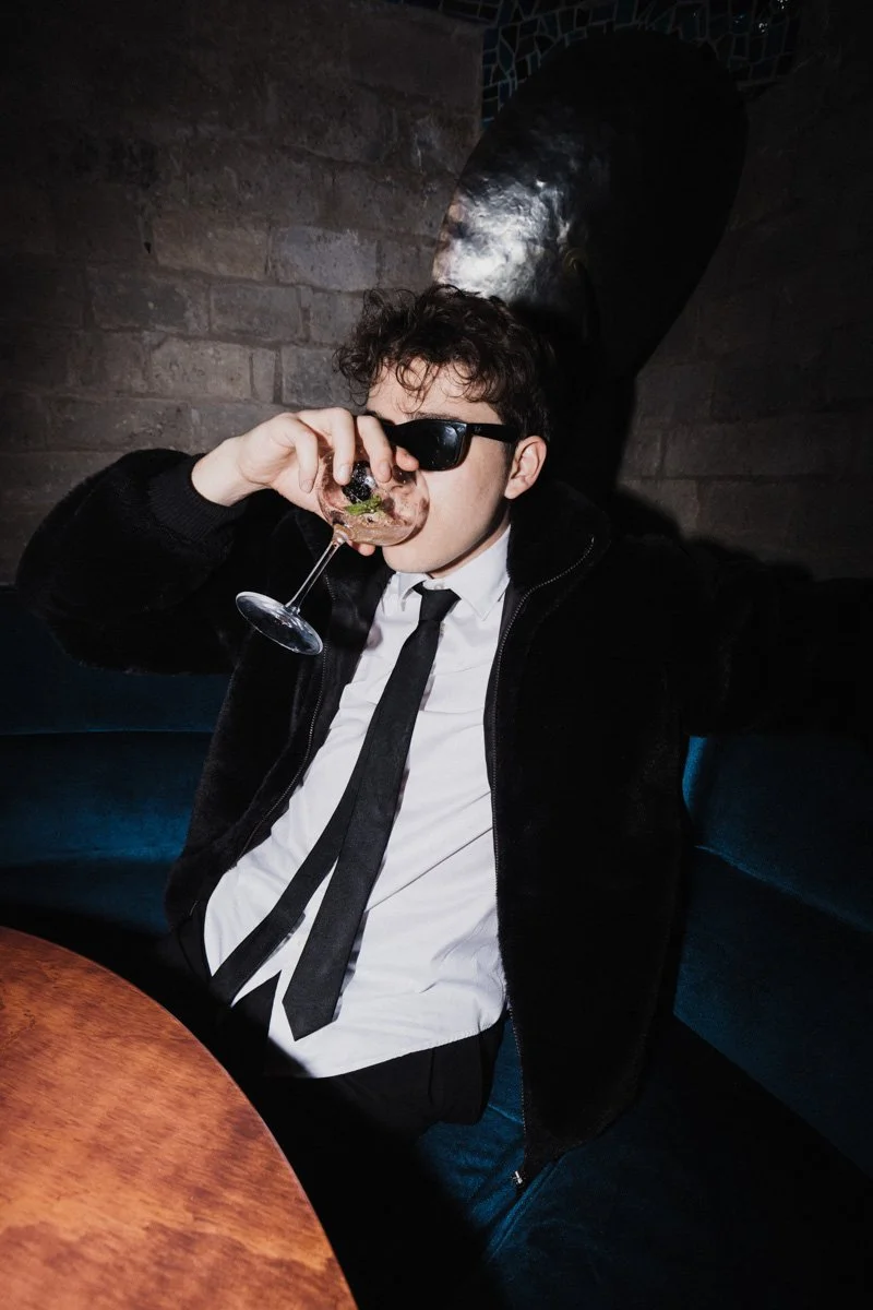 A young man with curly hair, wearing black sunglasses, a white shirt, a black tie, and a black jacket, drinks from a glass of wine while sitting in a dimly lit restaurant with a brick wall background.