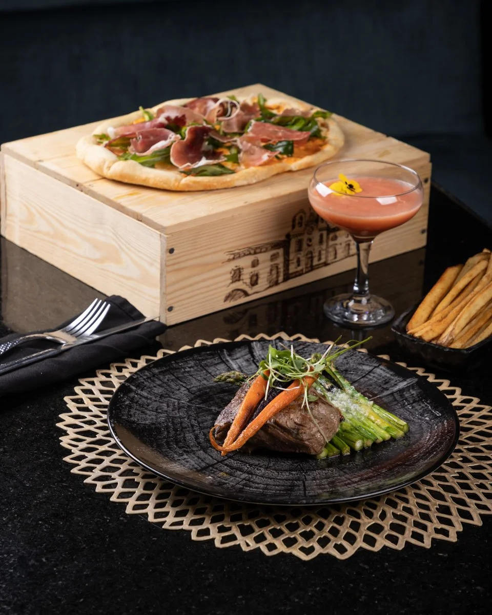 A gourmet meal with a seared steak, asparagus, and garnishes on a black plate, a wood-kept pizza with prosciutto, greenery, and cheese, a cocktail with a yellow flower, and a side of breadsticks. Food Photography in Mexico City by sergiogutierrezphot