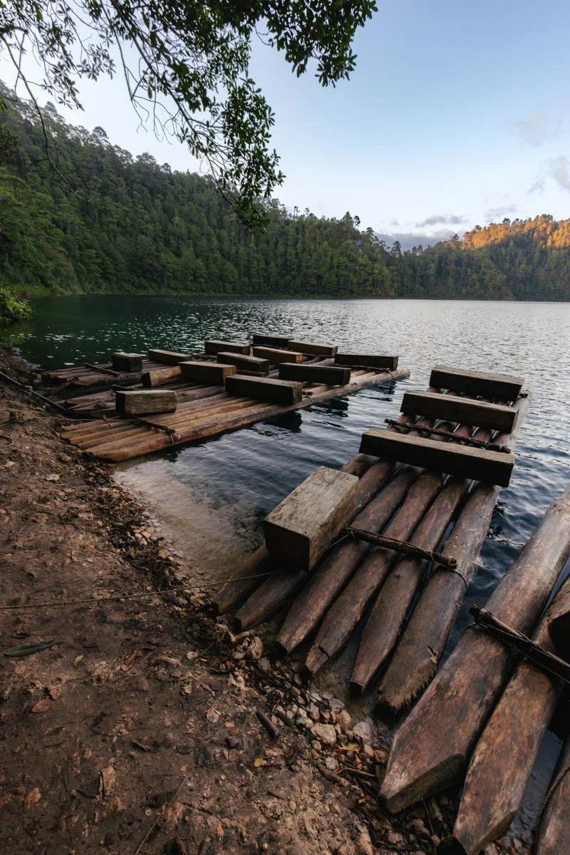Wooden floating dock on a lake surrounded by green forest, with rocky shore in the foreground and trees overhead. Lagunas de Montebello, Chiapas, Mexico. Travel Photography by Sergio Gutiérrez sergiogutierrezphoto.