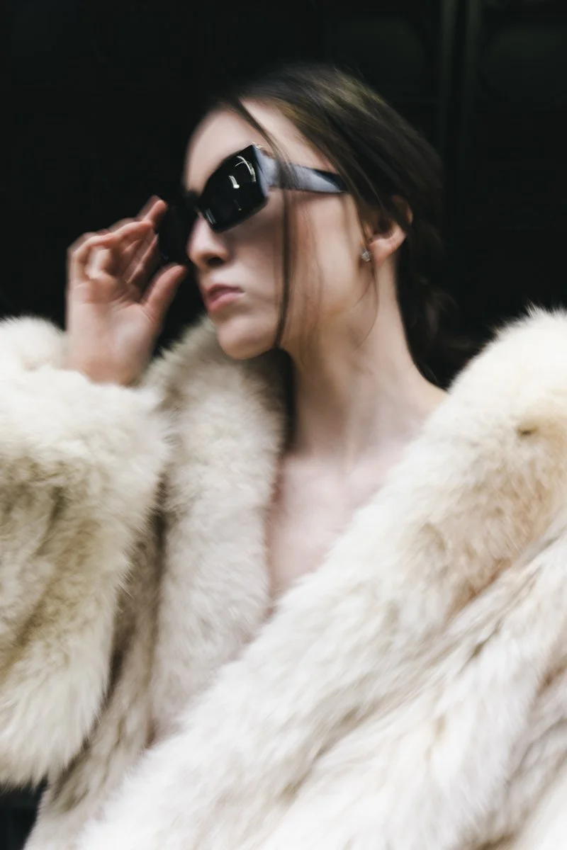 A woman wearing dark sunglasses and a white fur coat, adjusting her glasses with a serious expression. Fashion photography by sergiogutierrezphoto
