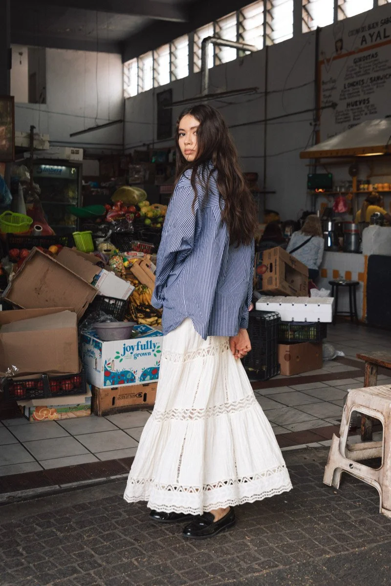 Young woman with long wavy hair, wearing a blue striped shirt and a white lace maxi skirt, standing in a market with produce and boxes in the background. Lifestlye photoshoot Mexico.