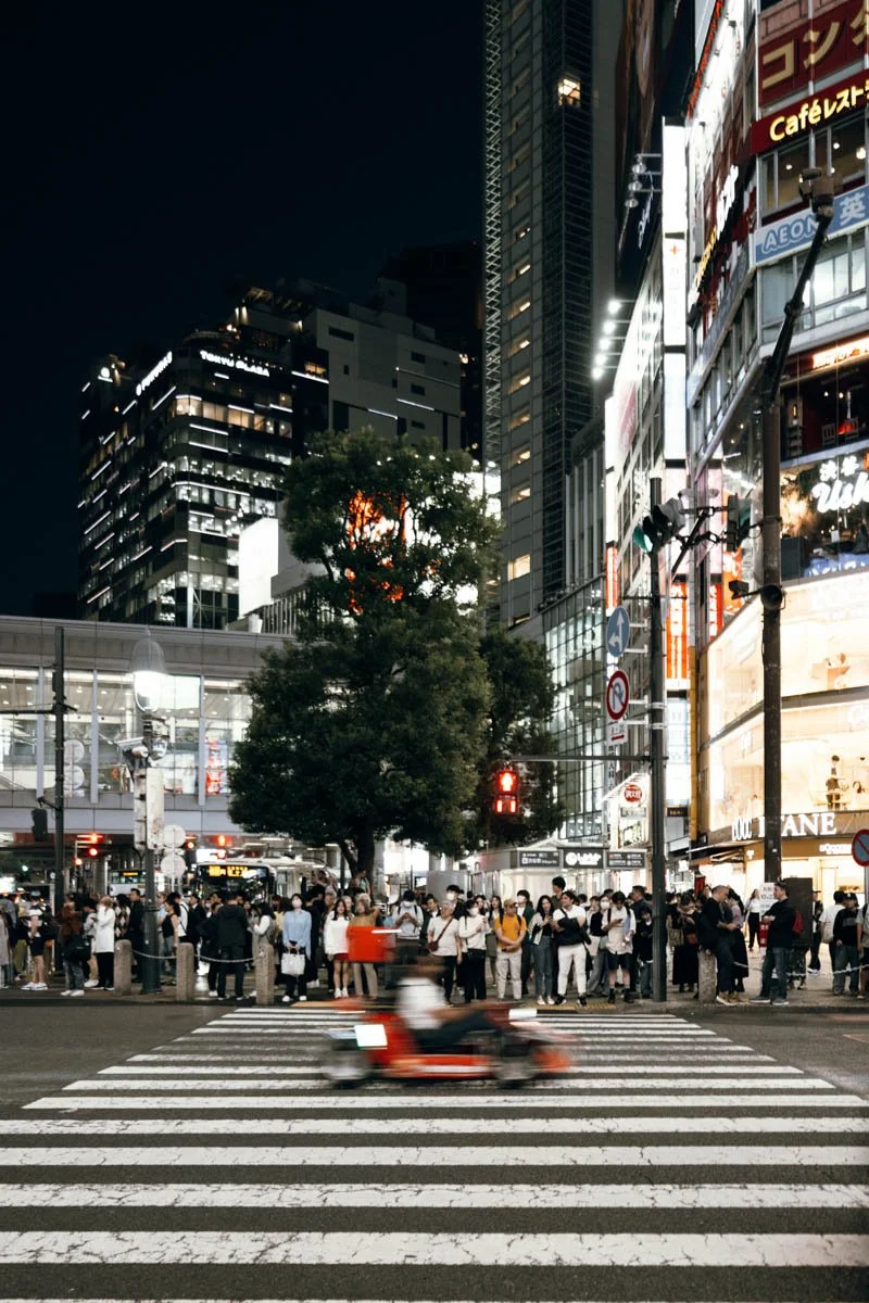 Nighttime city street scene with a crowd of pedestrians waiting to cross at a crosswalk, a person riding a blurred motorcycle in the foreground, tall illuminated buildings, and digital signage on the buildings. Shibuya Tokyo, Japan. 