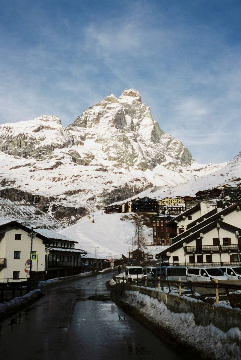 Snow-covered mountain with a small alpine village and parked vehicles in the foreground. Matterhorn Switzerland. Travel Film Photography By Sergio Gutiérrez sergiogutierrezphoto.
