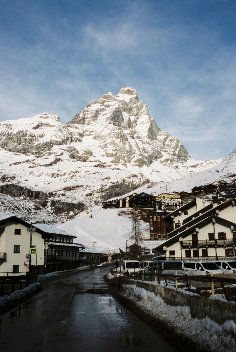 Snow-covered mountain with a small alpine village and parked vehicles in the foreground. Matterhorn Switzerland. Travel Film Photography By Sergio Gutiérrez sergiogutierrezphoto.
