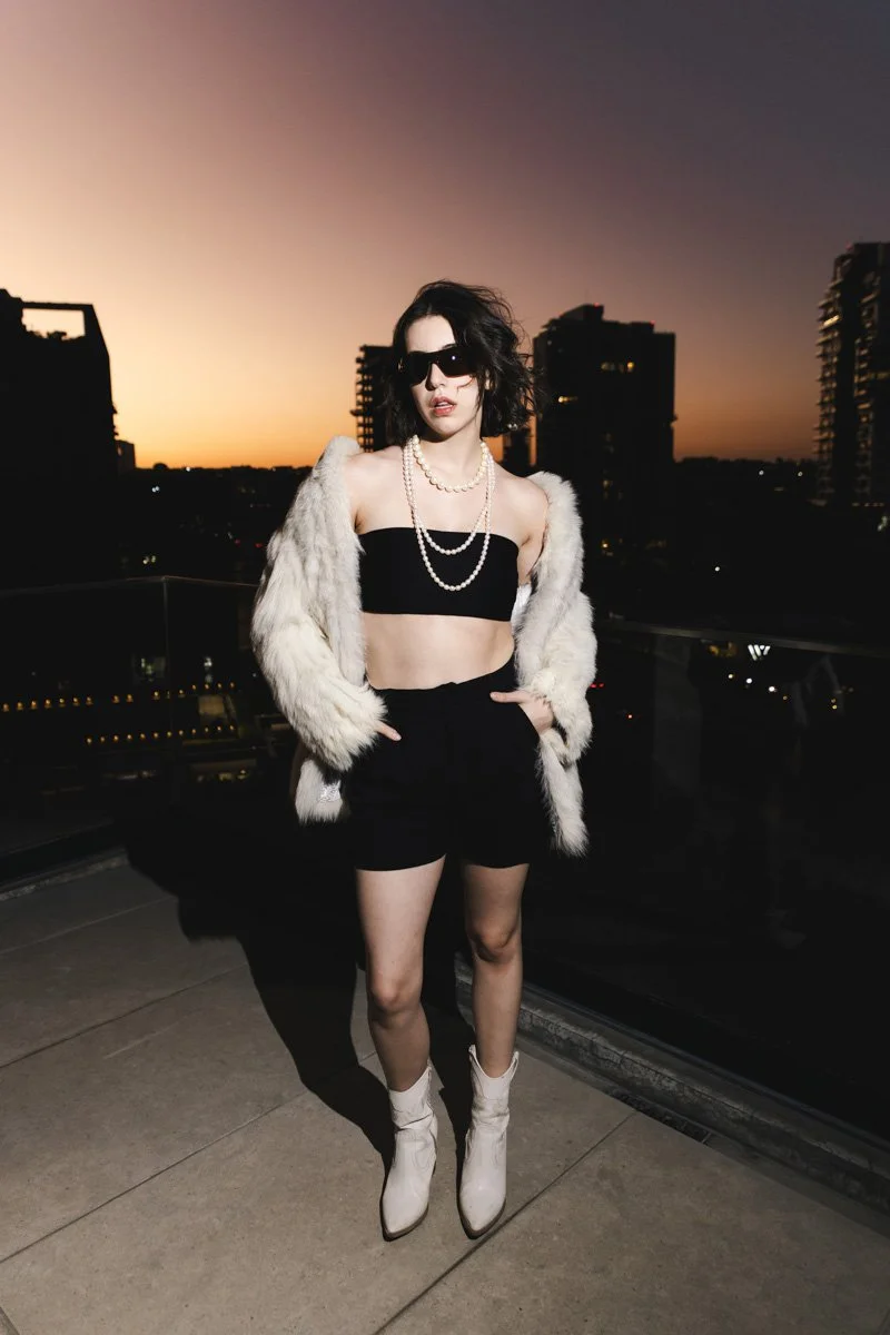 A young woman with short black hair and sunglasses standing on a rooftop during sunset, wearing a black strapless top, black shorts, white heeled boots, a white faux fur jacket, and pearl jewelry. Editorial photoshoot in Mexico.