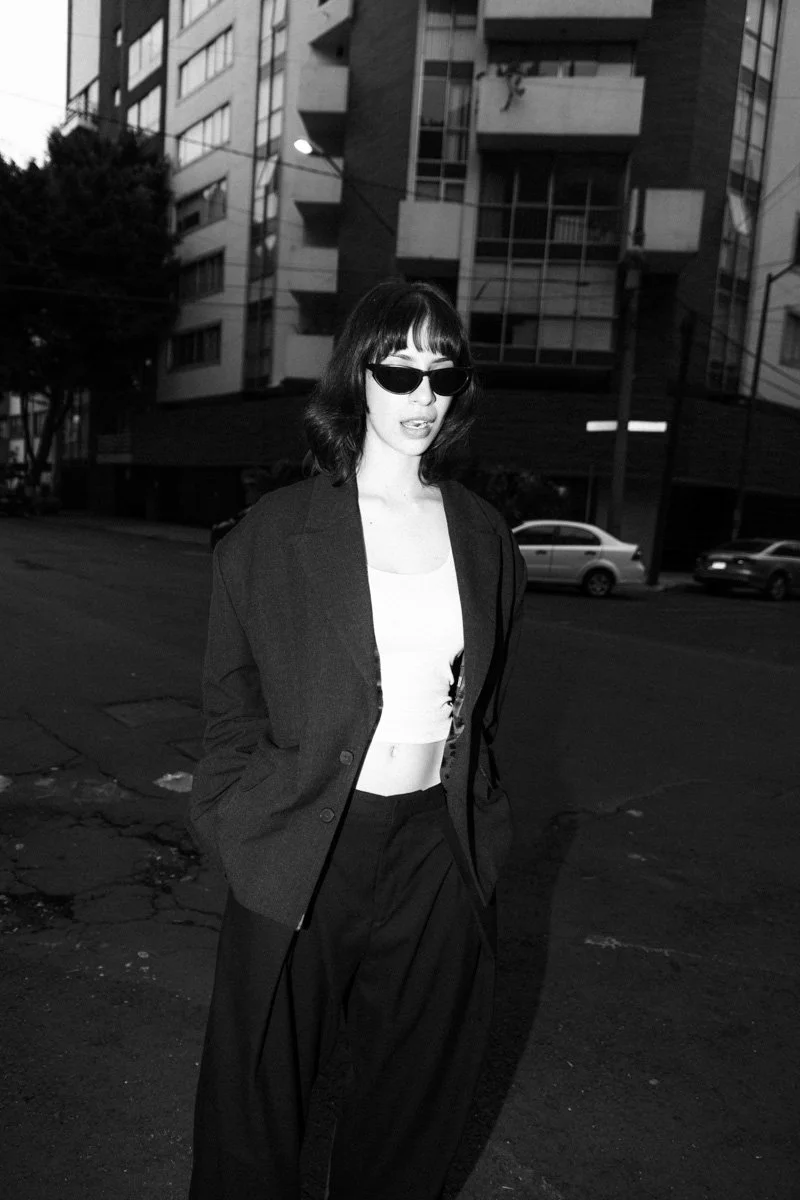 A woman with dark hair, wearing sunglasses, a white crop top, a dark blazer, and loose dark pants, standing on an urban street with tall apartment buildings and parked cars in the background, in black and white.