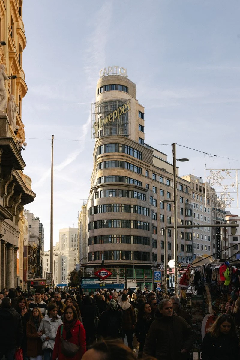 Crowded city street scene with a tall, curved building in the background that has a "Capitol" sign at the top and a "Schweppes" sign on the side. Madrid Spain travel photography by sergiogutierrezphoto