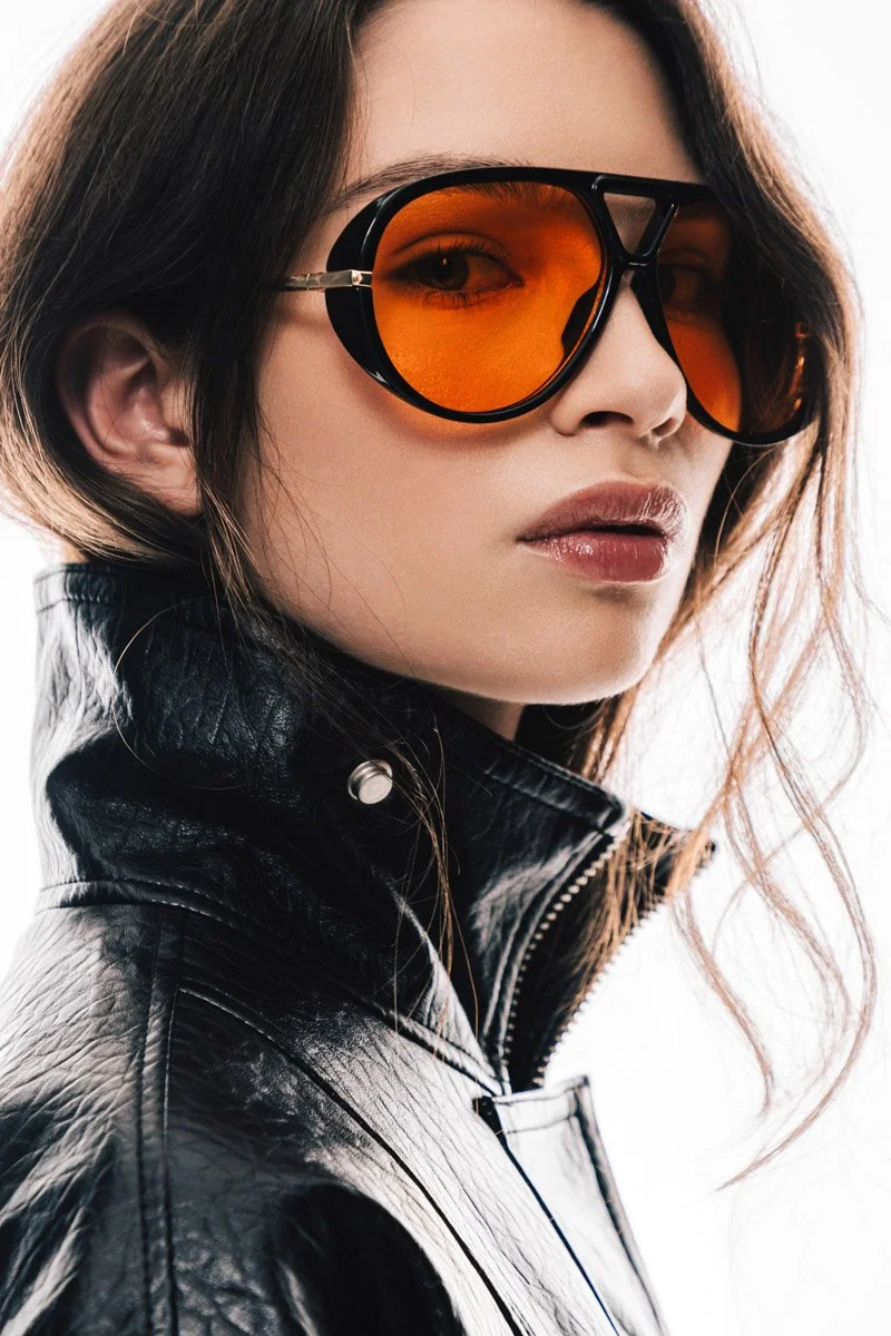 A woman with wavy brown hair wearing large black sunglasses with orange lenses and a black leather jacket. Commercial Fashion Photography. By Sergio Gutiérrez sergiogutierrezphoto. Mexico City