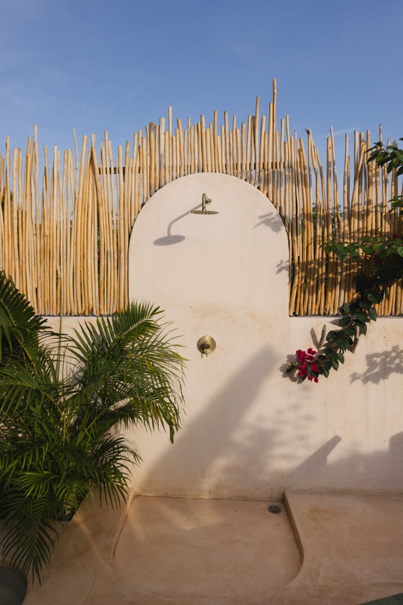 Outdoor outdoor shower with bamboo fencing, plants, and a white wall, under a blue sky. hotel photography puerto escondido mexico by sergiogutierrezphoto