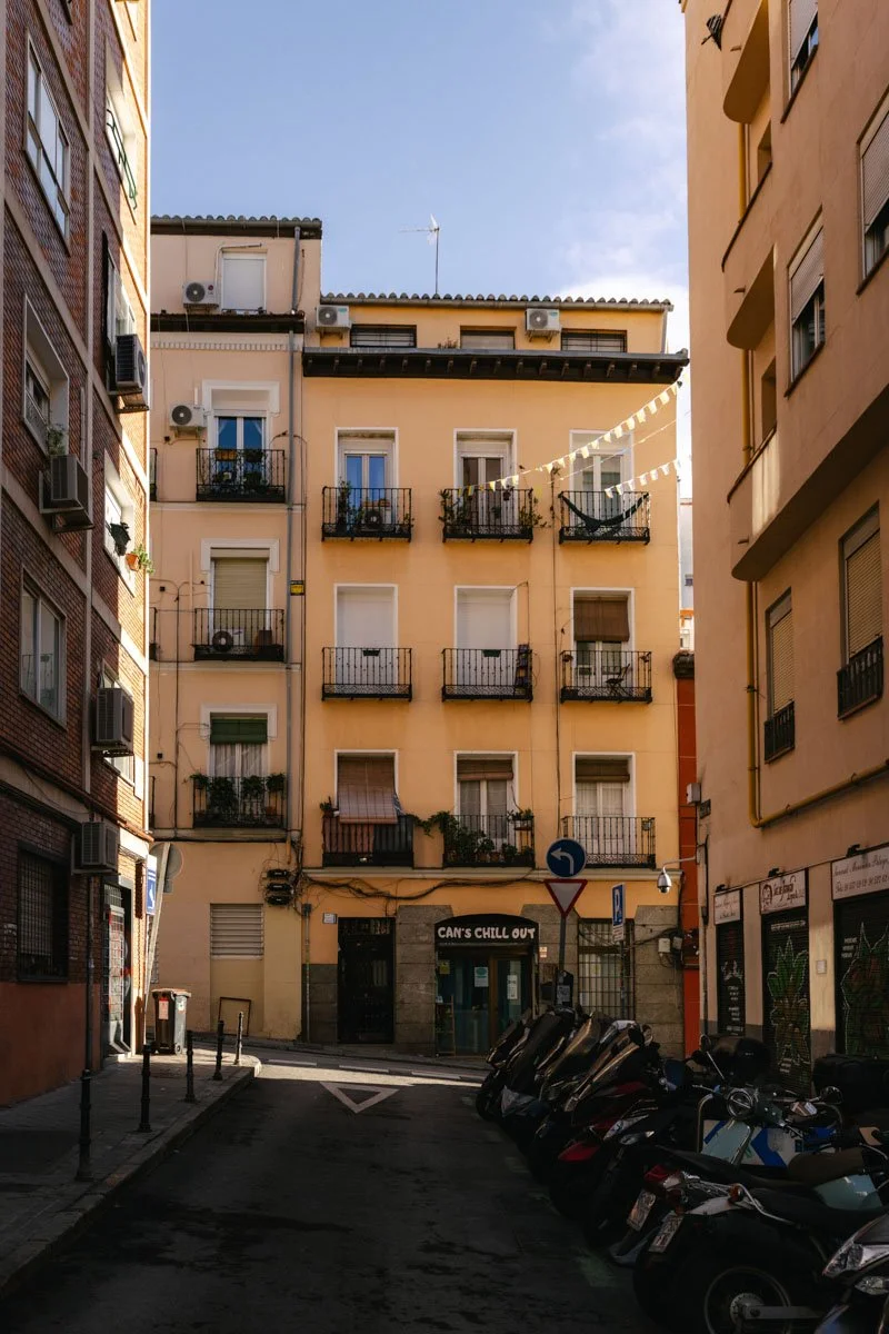 A narrow city street with multi-story buildings on both sides, parked motorcycles along the right curb, and a small business with a sign reading "CAN'S CHILL OUT" at the ground level. Madrid Lavapies travel photography by sergiogutierrezphoto