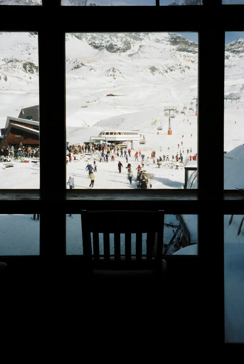 Snowy ski resort scene viewed through a window, with skiers and snowboarders on the slopes, ski lifts in operation, and a large building at the base. Travel Film Photography by Sergio Gutiérrez sergiogutierrezphoto.