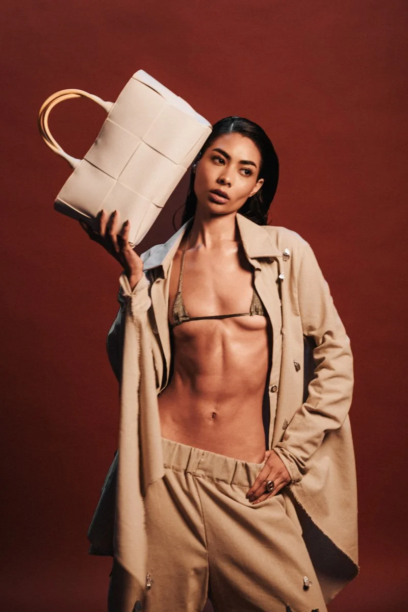 A woman with long dark hair, wearing beige pants and an open beige coat, holds a large cream-colored handbag up to her head. She is revealing her toned abdominal muscles and is wearing a small bikini top against a solid reddish-brown background. 
