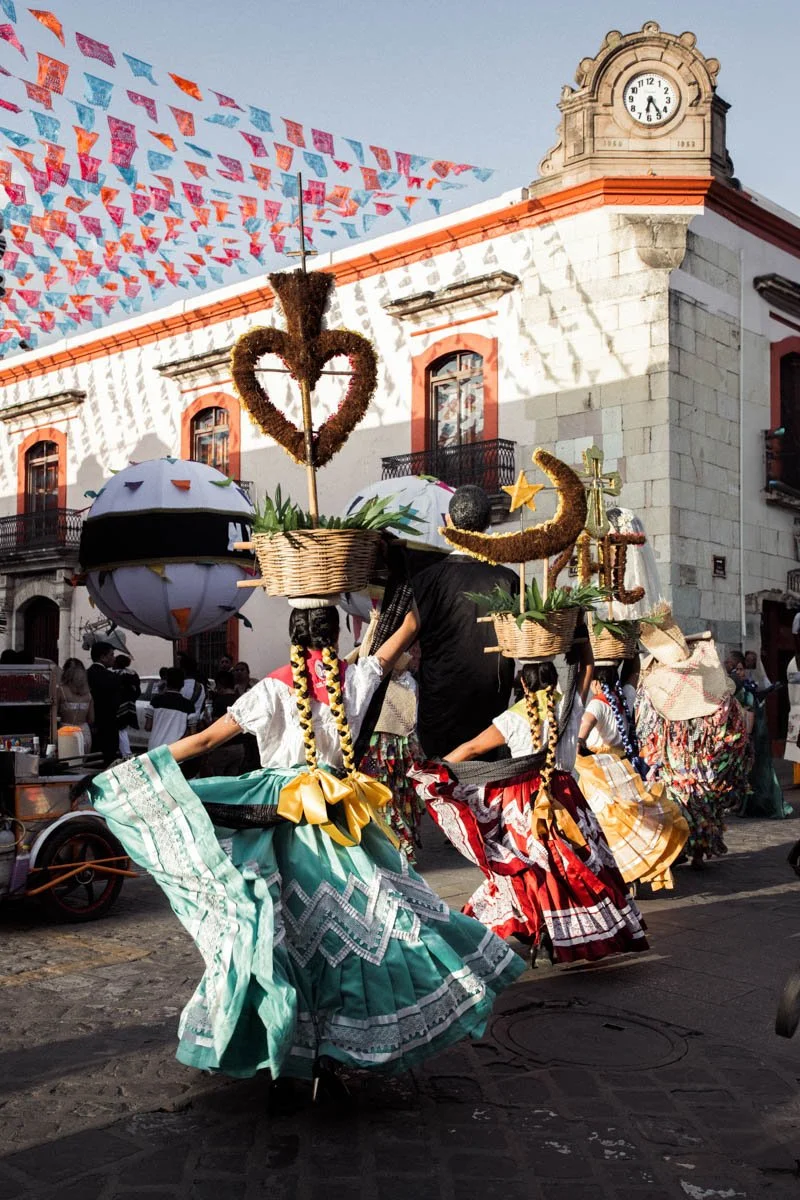 People participating in a traditional parade wearing colorful dresses, with headpieces decorated with baskets, hearts, and moons, walking past a historic building with a clock tower and colorful papel picado banners overhead. Oaxaca Mexico Wedding.