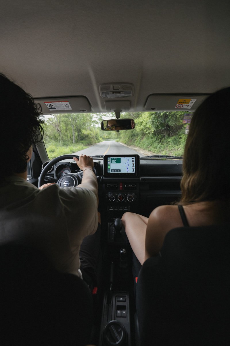 View from inside a car showing two passengers, one driving and one sitting on the passenger side. The car is on a narrow, winding road surrounded by green trees. Puerto Escondido Oaxaca Mexico.