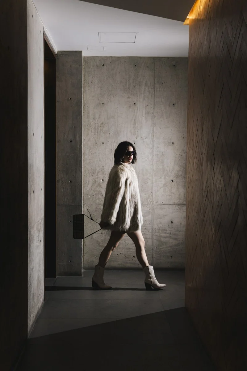 A woman in sunglasses wearing a fur coat and high-heeled boots walks through a modern, minimalistic interior with concrete and wooden walls. Fashion photography in Mexico.