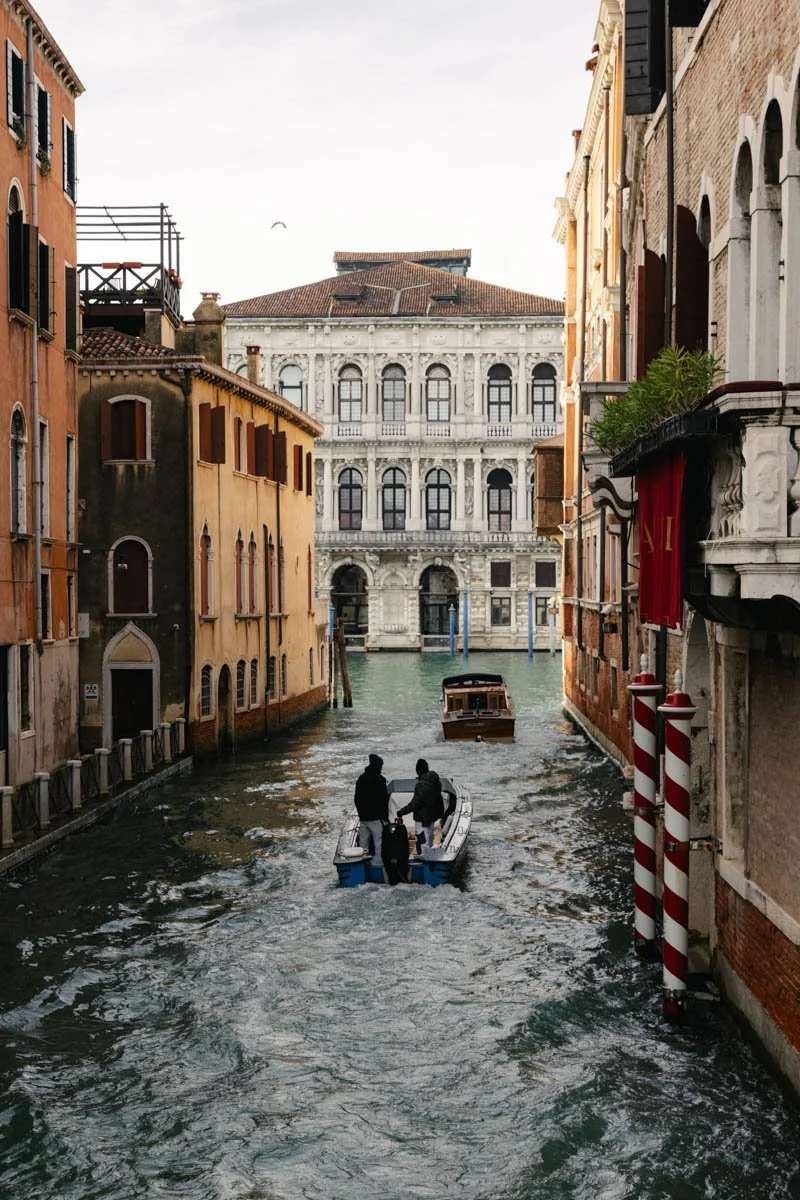 A canal in Venice, Italy, with buildings on both sides and two small boats carrying passengers. Travel Photography By Sergio Gutiérrez sergiogutierrezphoto.