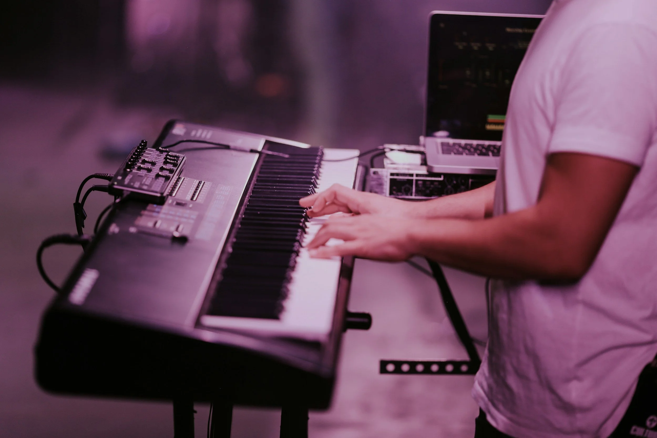 A person wearing a white t-shirt playing a keyboard synthesizer with a laptop nearby in a dimly lit room with purple lighting.