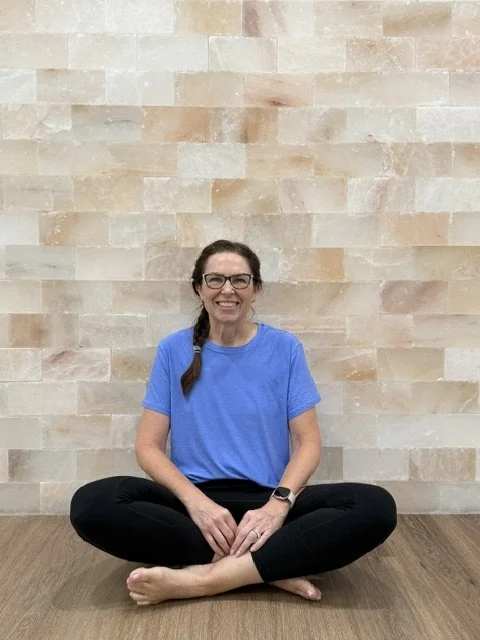 A woman sitting cross-legged on the floor in front of a beige stone wall, smiling, wearing glasses, a blue t-shirt, and black pants.