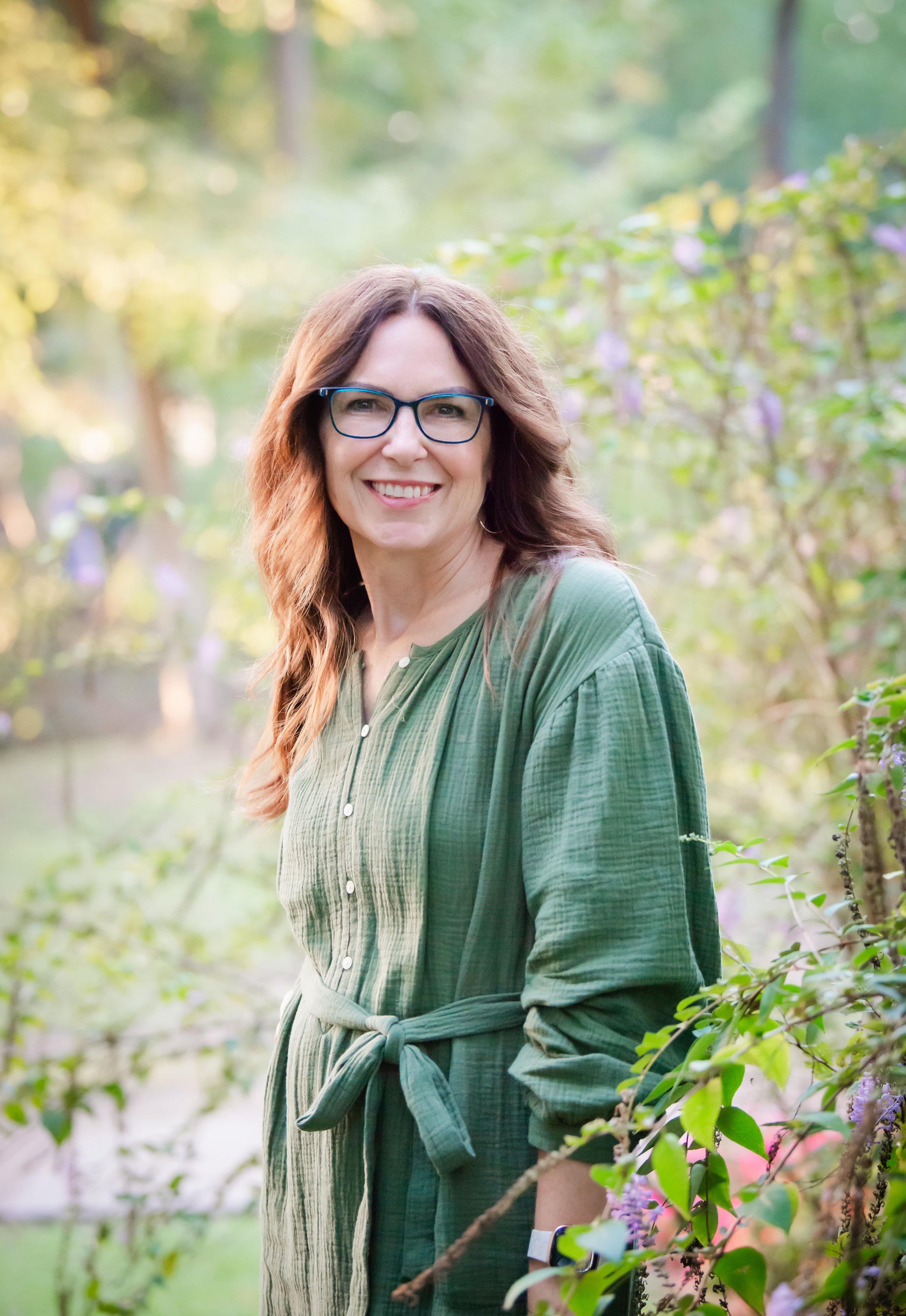 A woman with glasses and long, wavy hair smiling outdoors among green foliage and purple flowers, wearing a green dress with a waist tie.