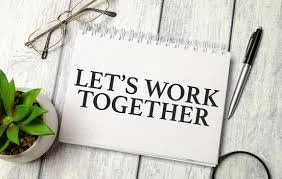 Notepad with the phrase 'Let's Work Together' written on it, next to glasses, a pen, and a small potted plant on a white wooden surface.