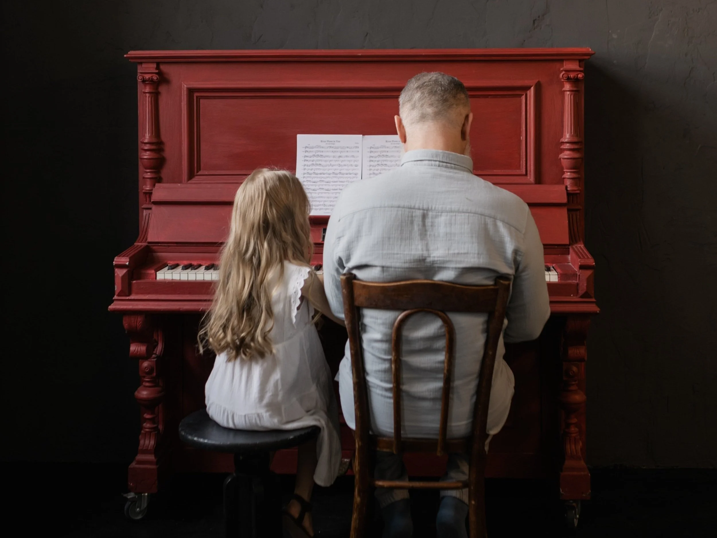 Child with long blond hair and adult man seated together at a red piano