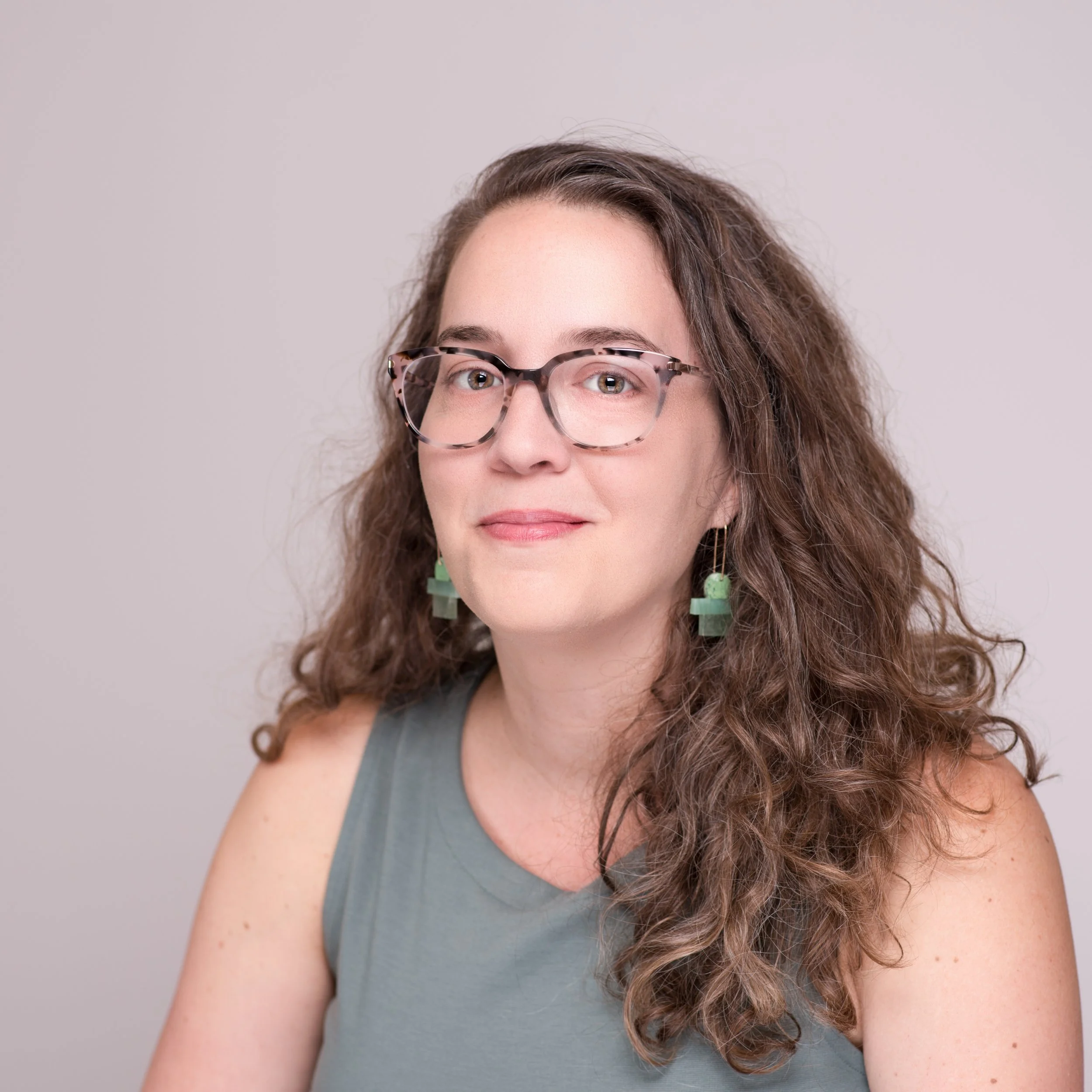 Close-up of a woman with long curly hair, wearing round glasses with a tortoise shell pattern, and a sleeveless gray-green top.