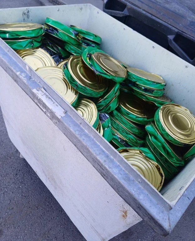 Metallinen laatikko täynnä puristettuja säilyketölkkejä, vihreitä ja kultaisia.