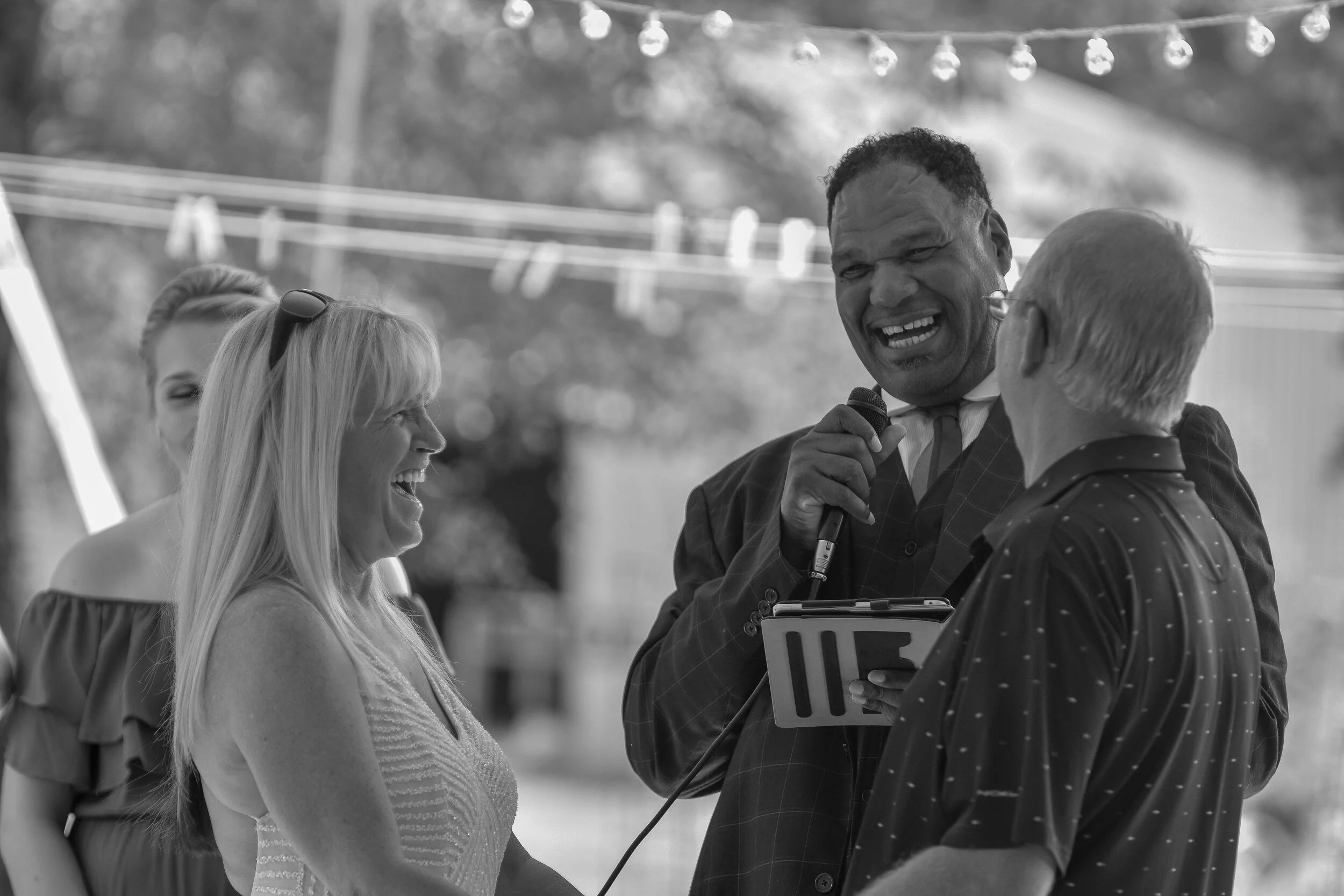 A group of people gathering outdoors, with one man holding a microphone and speaking, while others smile and listen, at a wedding.
