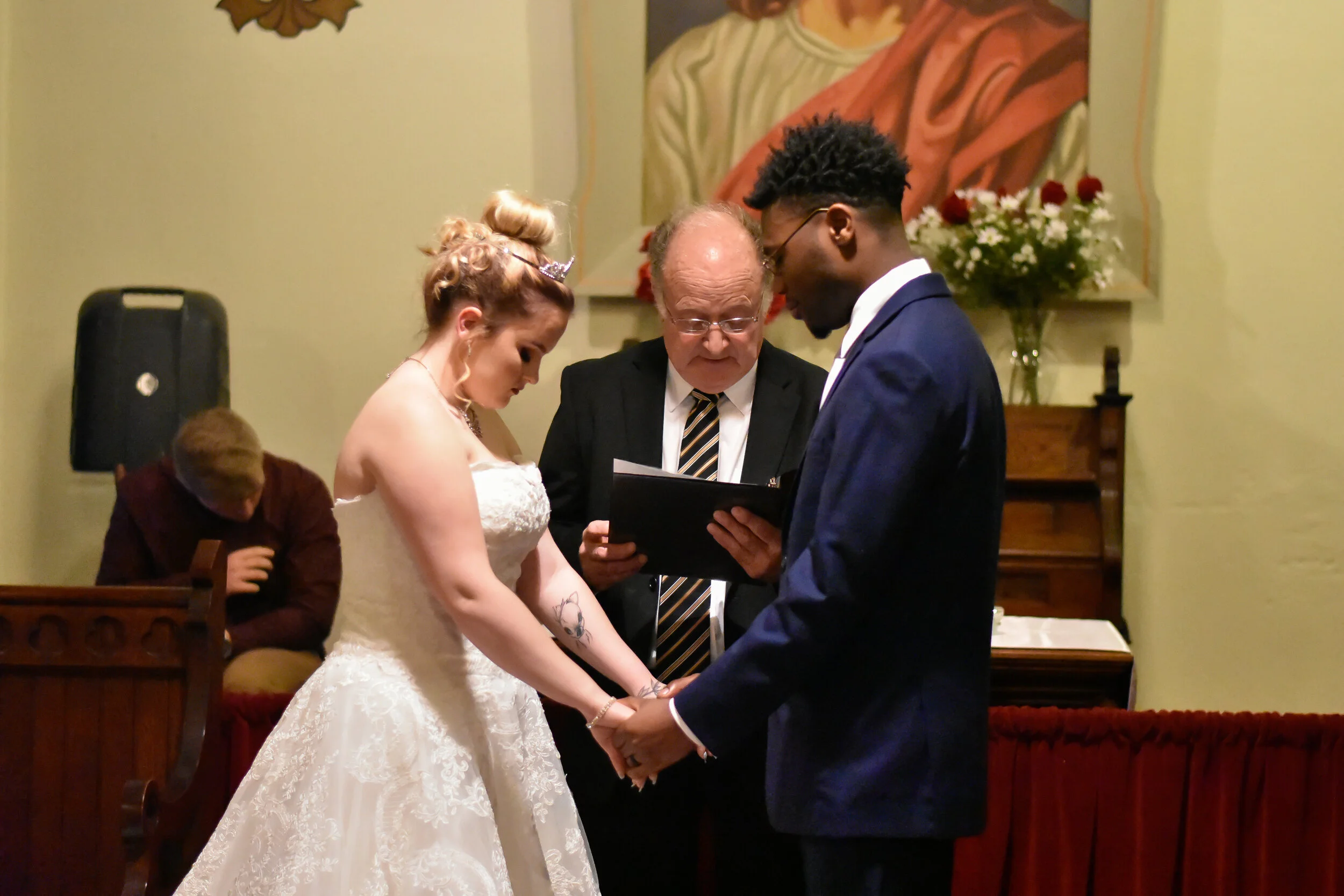 A couple getting married, holding hands during their wedding ceremony with an officiant reading from a book. The bride wears a white lace gown with tattoos visible on her arms, and the groom wears a navy blue suit. A person in a brown jacket is seate