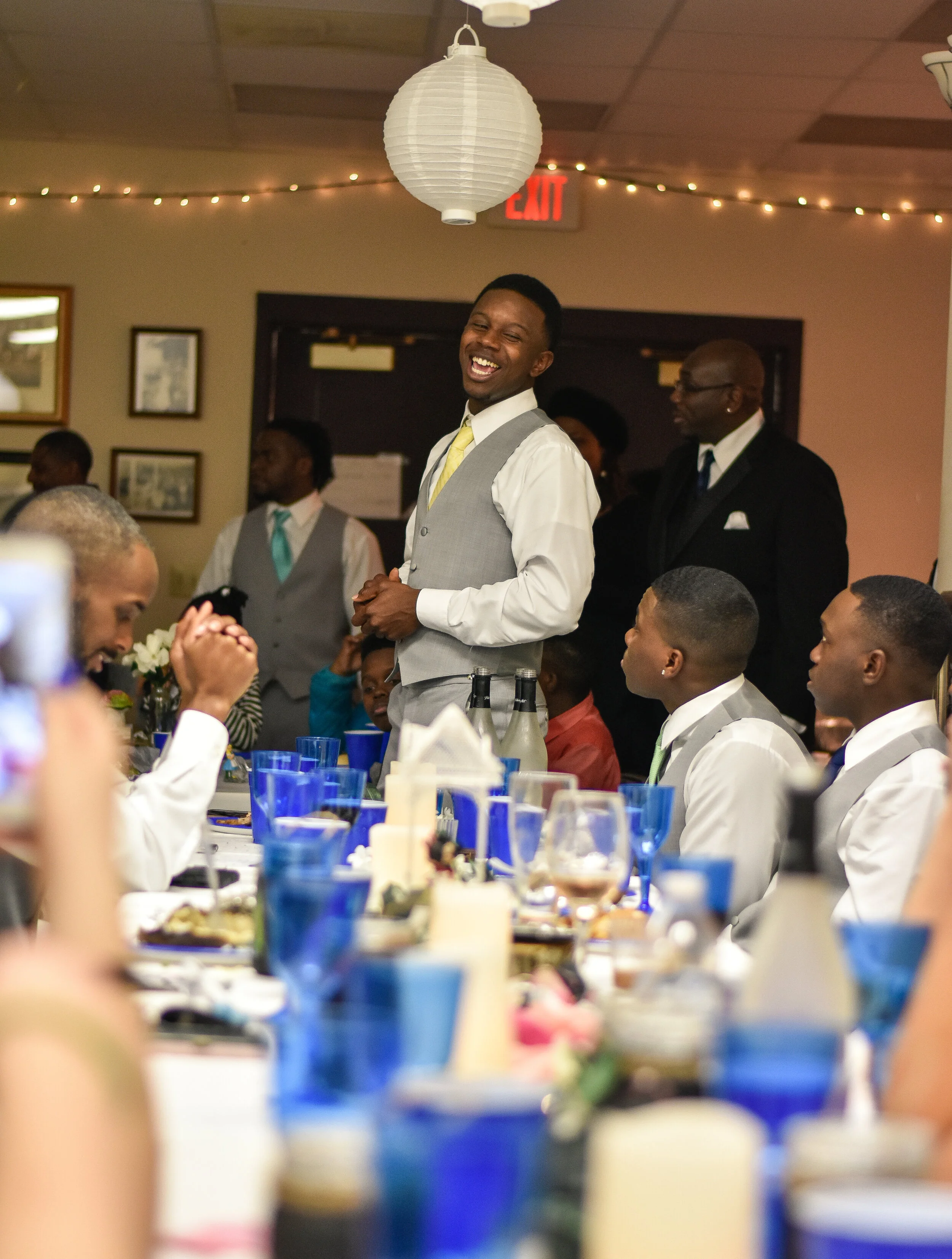 A man in a gray vest and yellow tie is laughing and standing at a long table during a celebration or wedding, surrounded by seated guests and a few people standing in the background, in a decorated banquet hall.