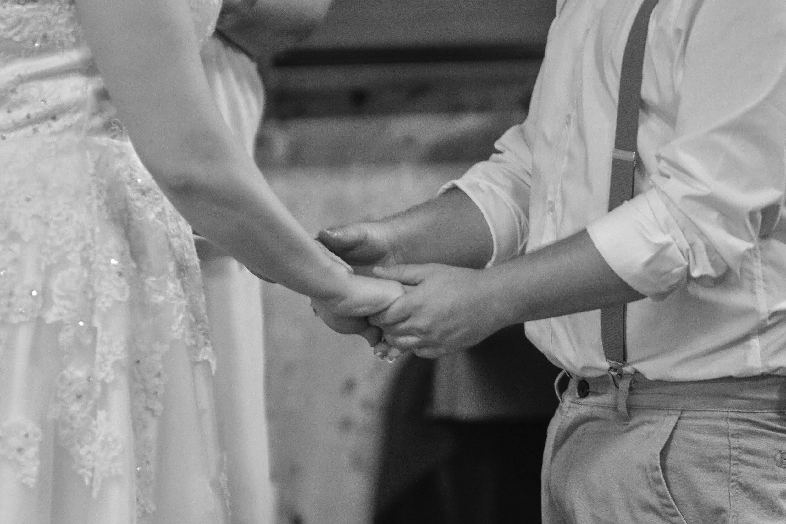 A couple holding hands, the woman wearing a wedding dress and the man dressed in a shirt with suspenders.