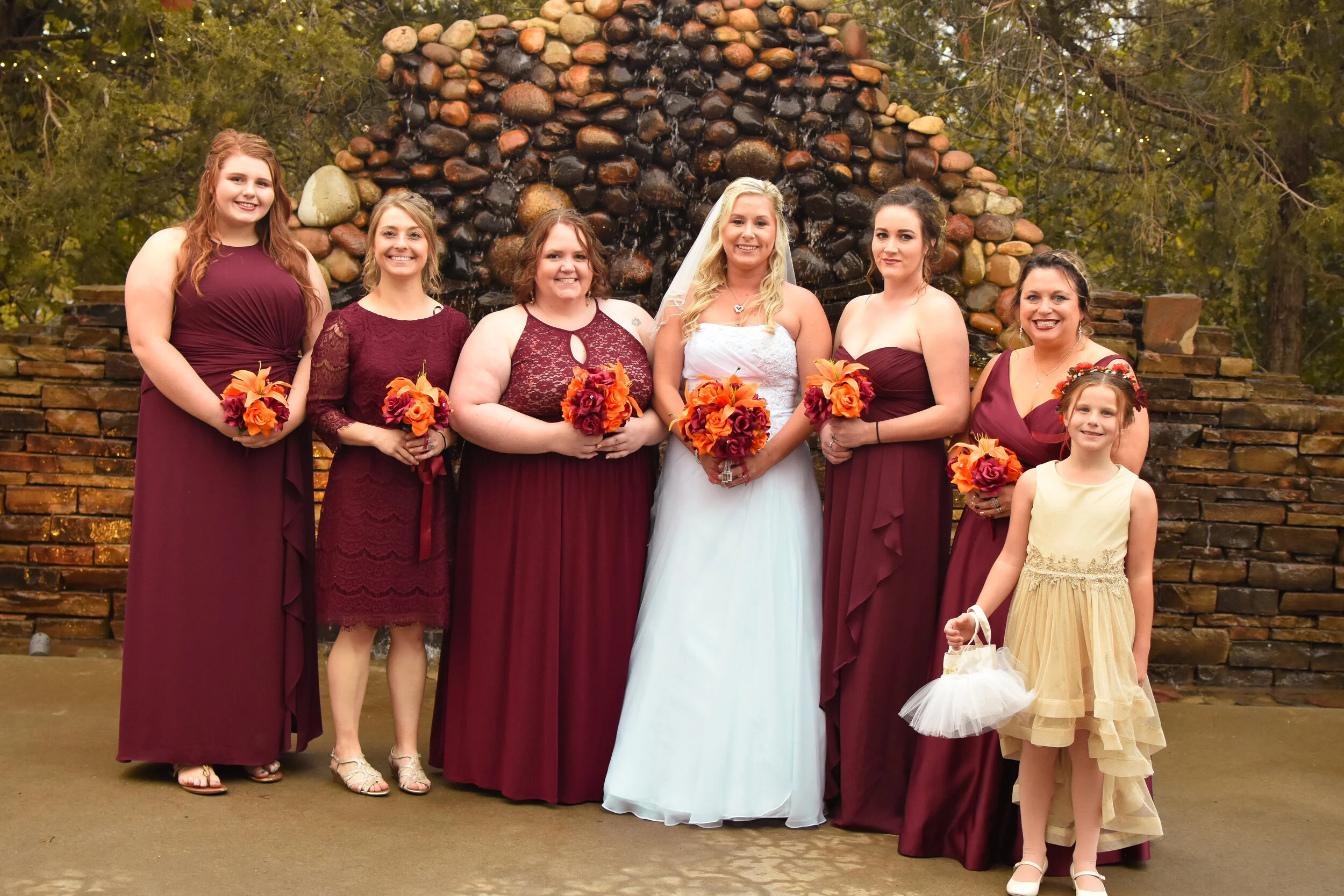 Group of women and a girl at a wedding, standing in front of a stone structure, holding bouquets with orange and red flowers. The bride is in the center wearing a white wedding dress and veil. Bridesmaids are in burgundy dresses, and a young girl is 