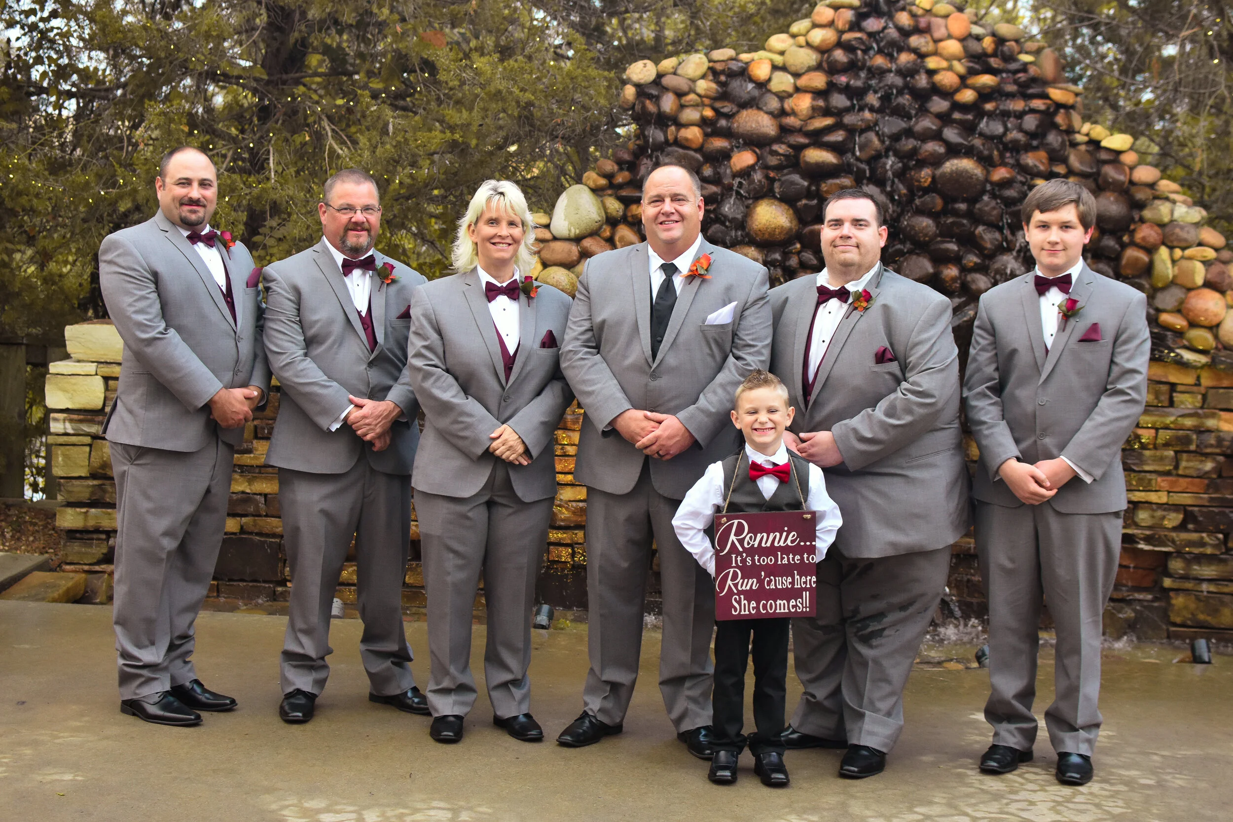 A wedding party with seven people, including six adults and one child, standing outdoors in front of a rock fountain. They are dressed in matching gray suits with red bows, and the child is holding a sign that says 'Ronnie... It's too late to run 'ca