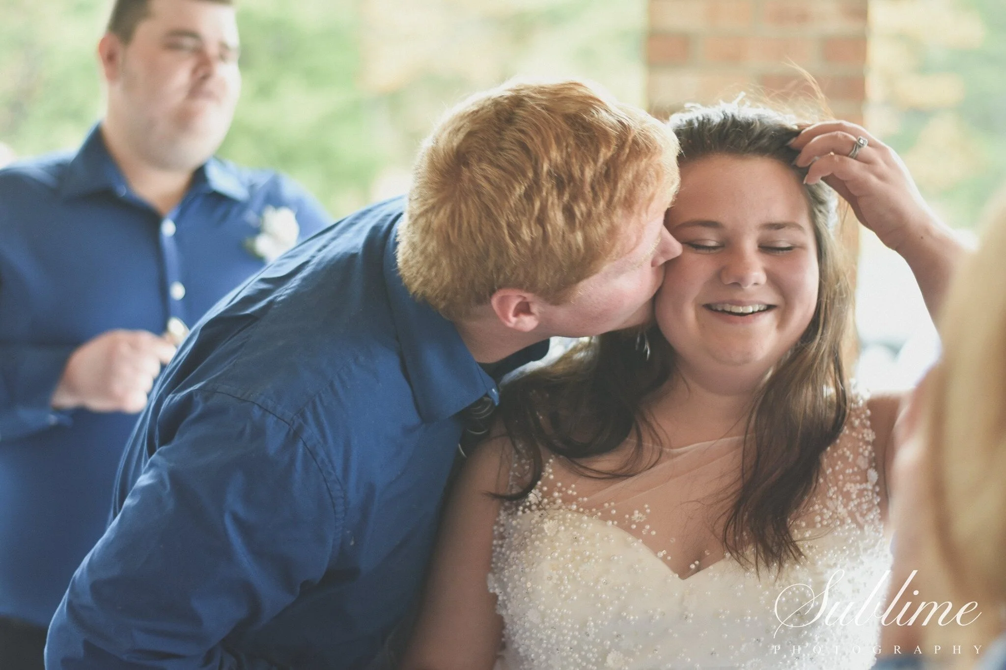 A man with red hair kissing a smiling woman with brown hair on the cheek at a wedding reception. The woman is wearing a white dress with beading, and a man with a blue shirt is in the background.