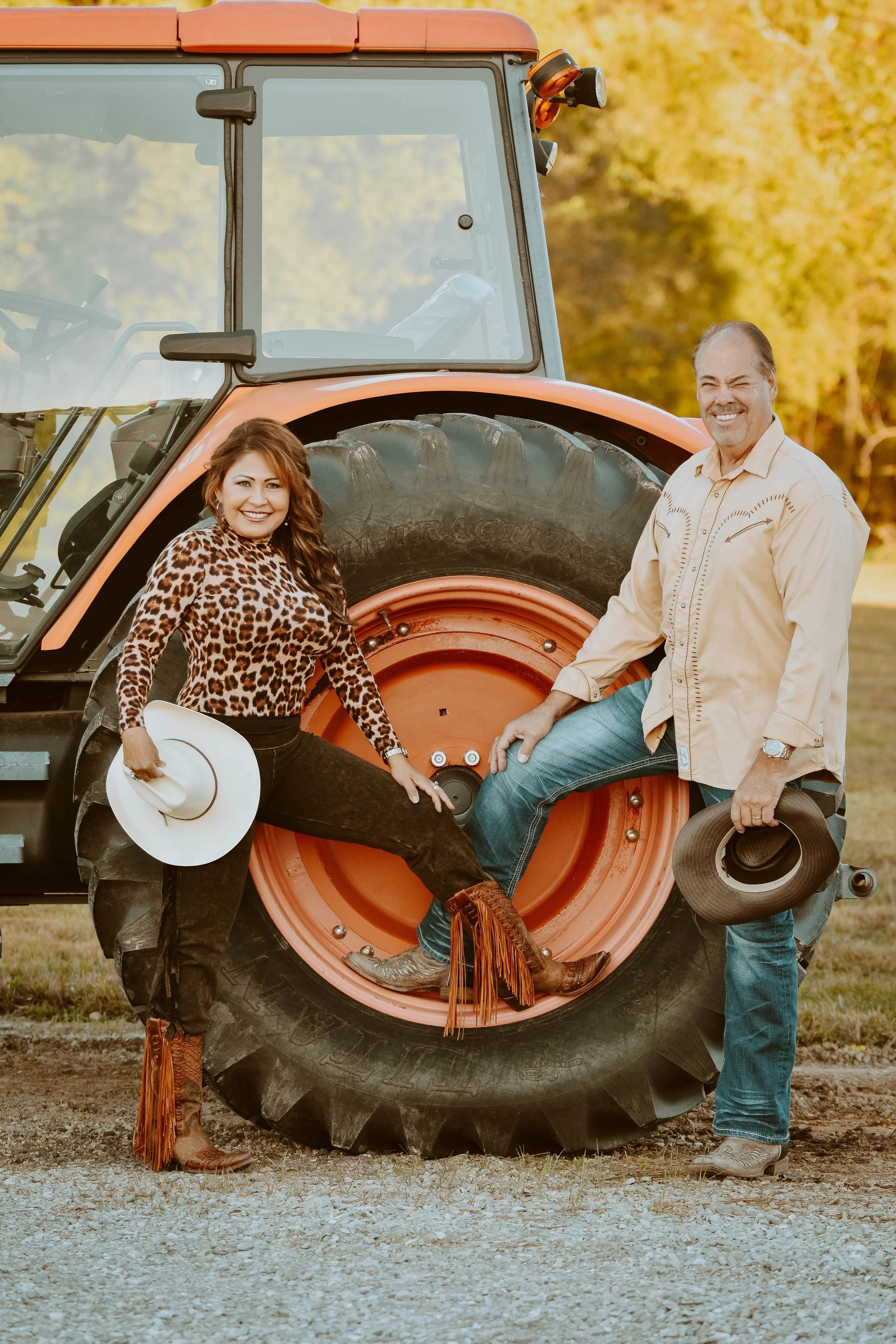 A woman and a man posing next to a large tractor tire, outdoors during sunset.