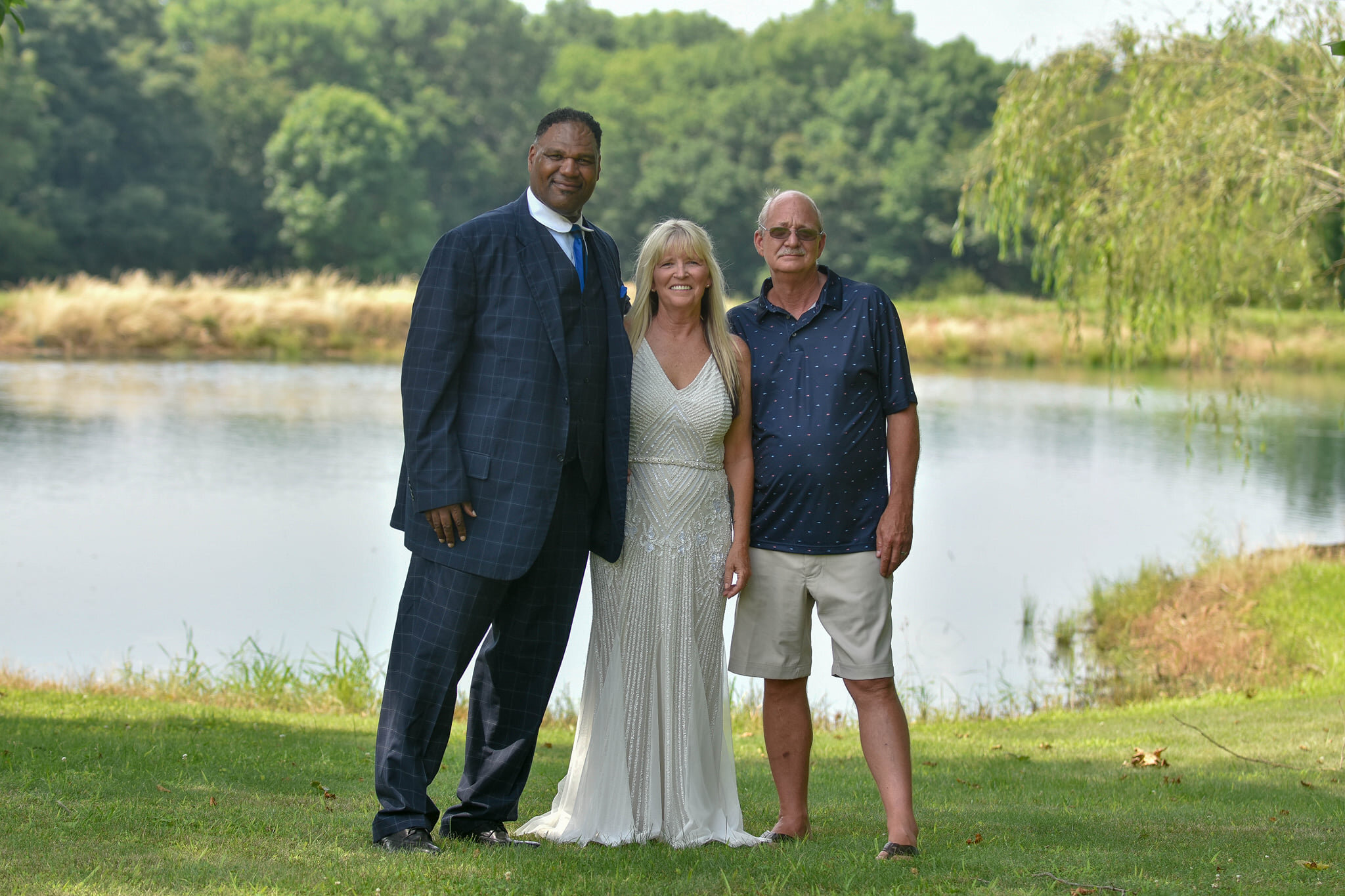 A man in a dark blue checked suit, a woman in a white dress, and a man in a navy polo and beige shorts standing by a lake with trees in the background during daytime.