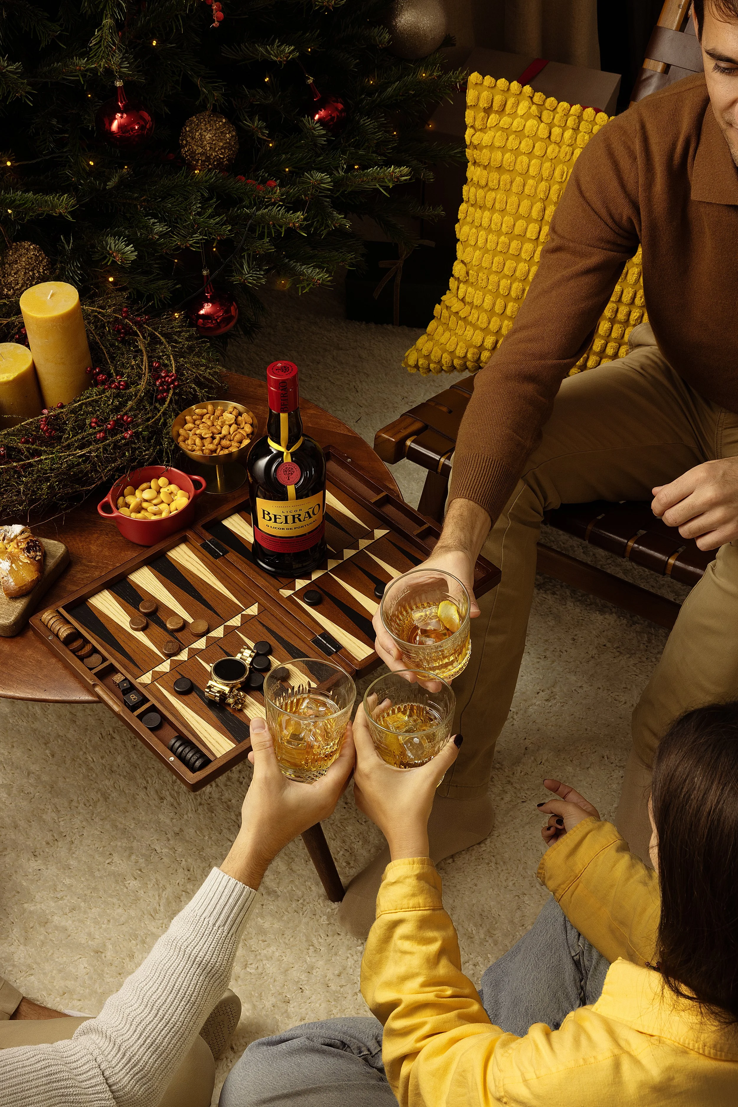 Reunião de pessoas jogando damas e brindando com drinks em uma sala decorada para o Natal, com uma árvore de Natal ao fundo.