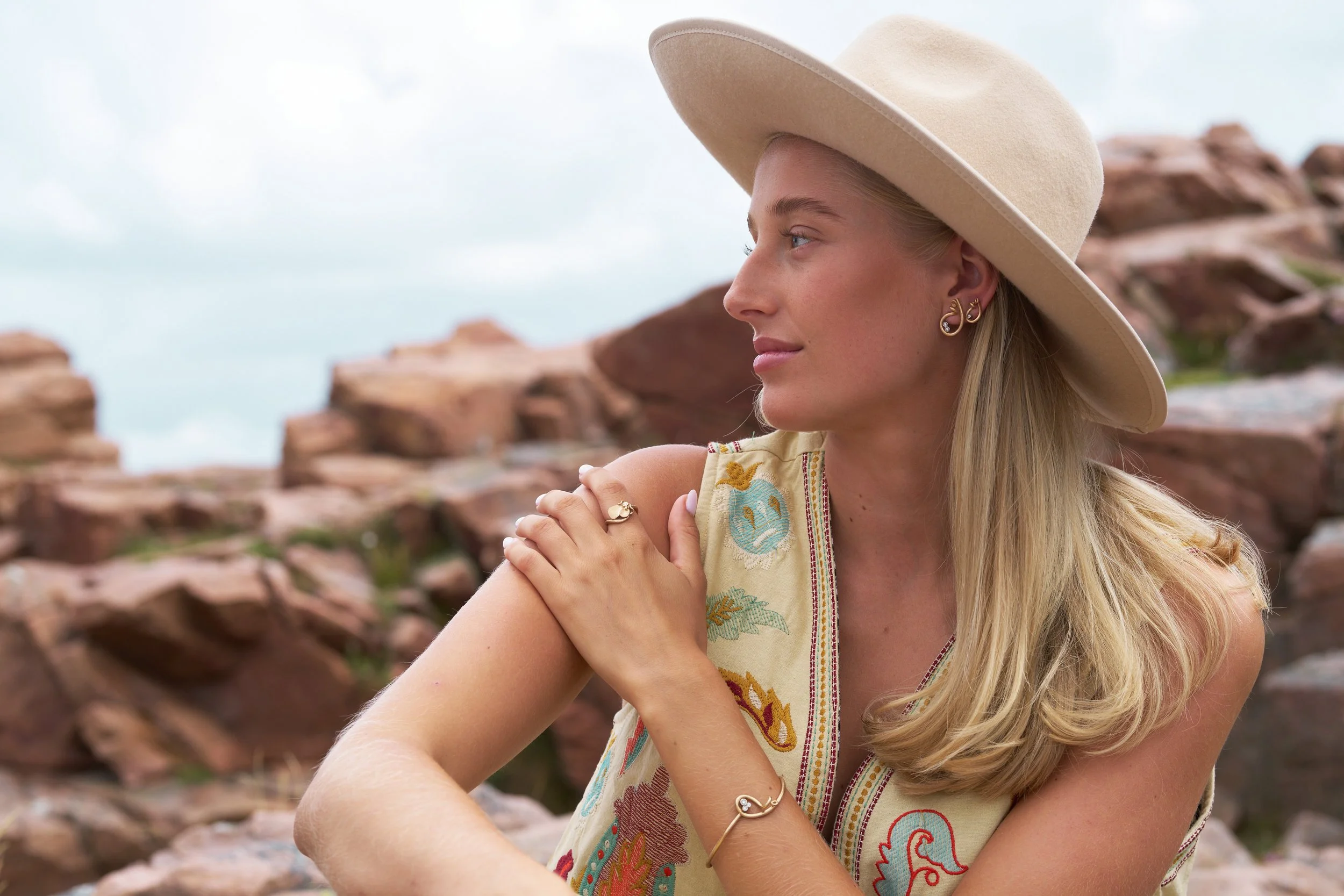 A woman with long blonde hair wearing a wide-brimmed beige hat and colorful embroidered sleeveless top, sitting outdoors with rocky terrain in the background.