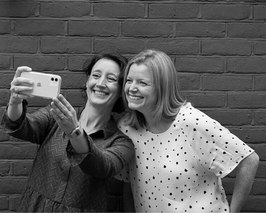 Julie + Sophie from Nutshell taking a selfie together in front of a brick wall, smiling and posing for the photo.