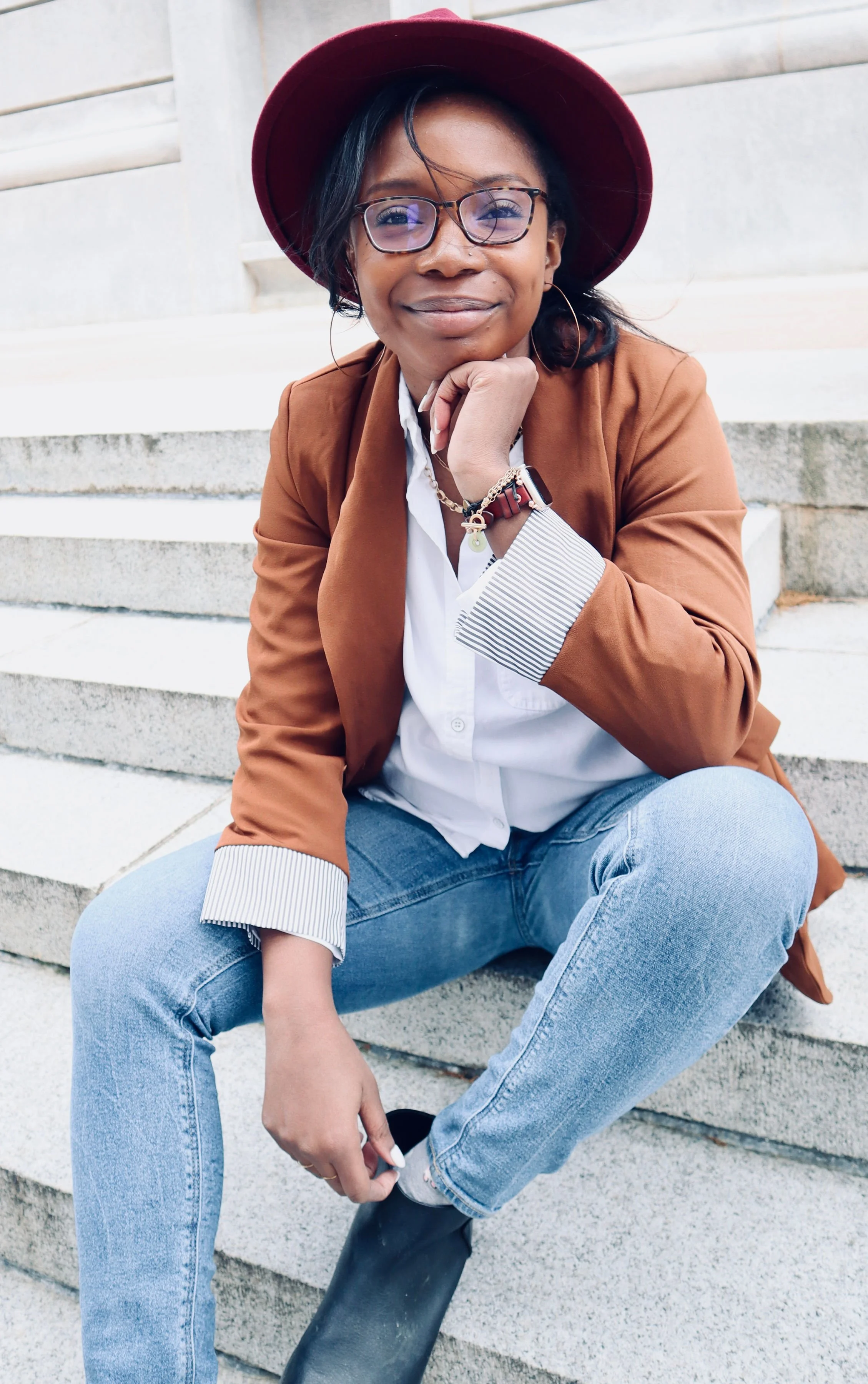 Erica who is a black woman smiles at the camera wearing a brown blazer, white blouse, blue jeans, and a maroon hat. She is wearing glasses and sitting outside on cement steps