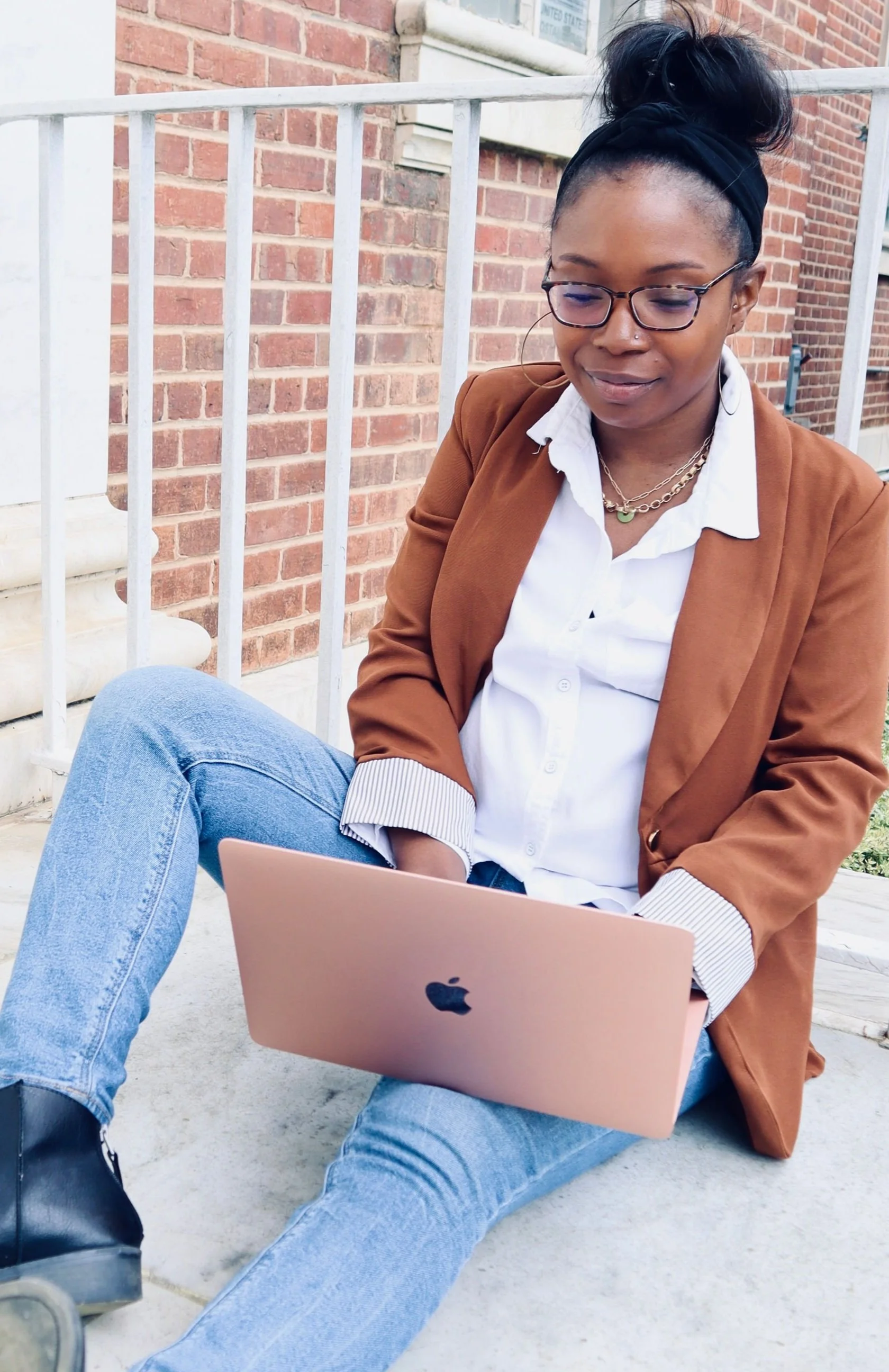 Erica sits outside typing on her laptop.. She is a black female wearing her hair in a top knot, wearing a brown blazer and blue jeans.
